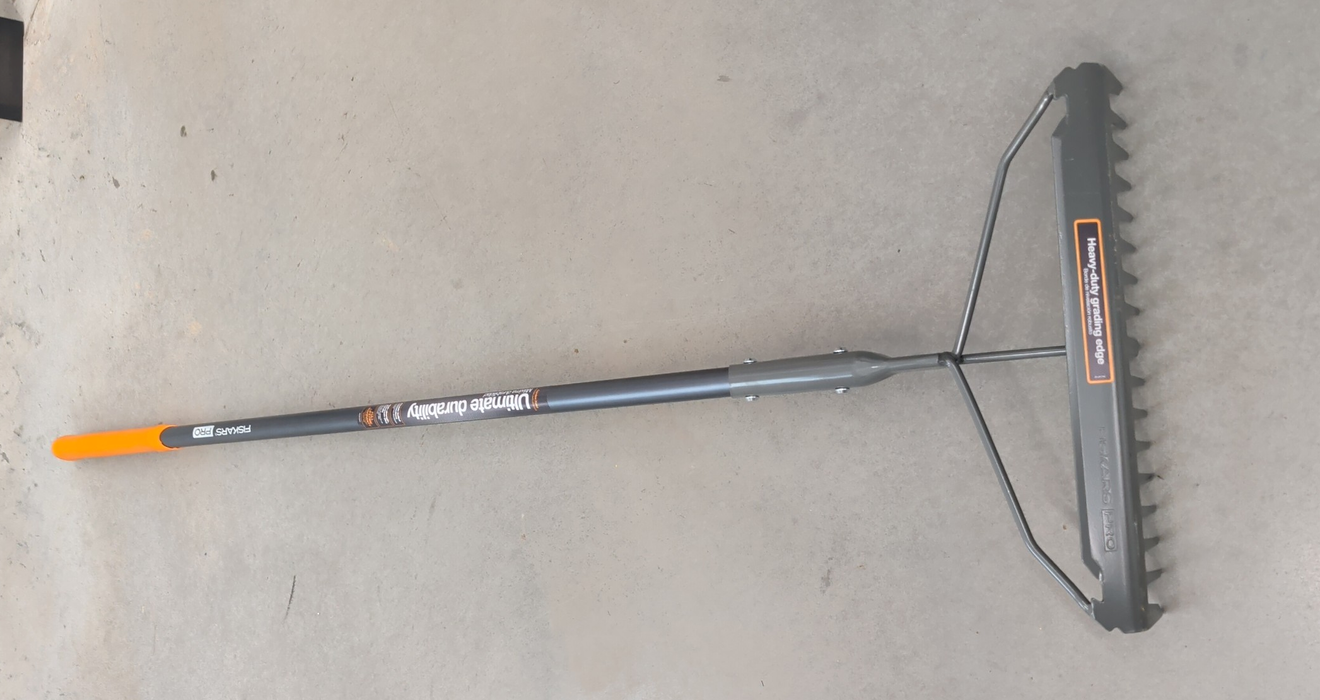 A Fiskars PRO garden rake featuring a dual-sided head with rigid teeth and a smooth grading edge, connected to a long grey handle with an orange grip.